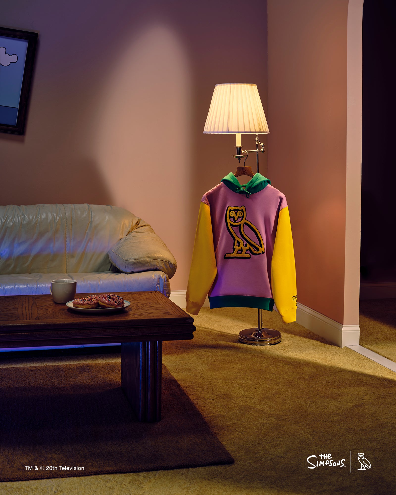 Purple and yellow October's Very Own hoodie on a mannequin in a dimly lit room with a couch and lamp.