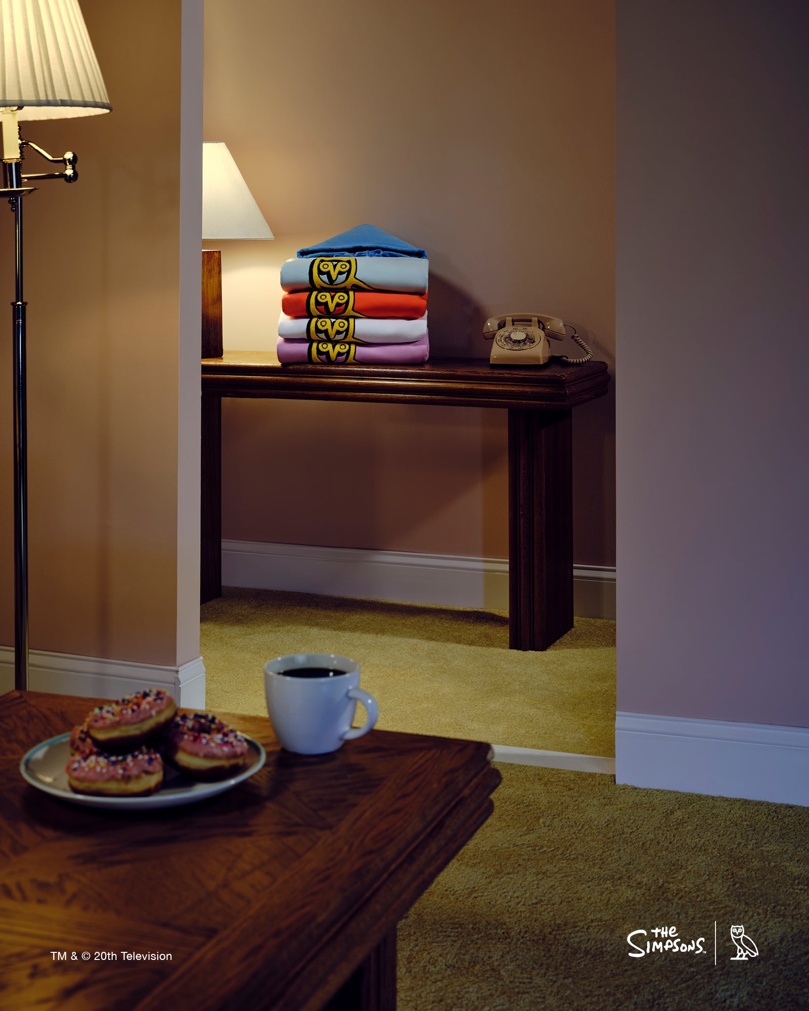 Stack of folded October's Very Own x The Simpsons sweaters on a wooden table with a cup and donuts in a room setting.