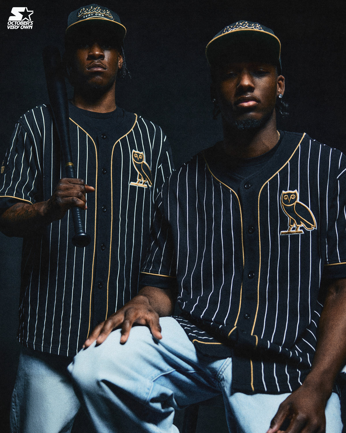 Two individuals wearing matching baseball jerseys with a October's Very Own logo, holding bats against a dark background.