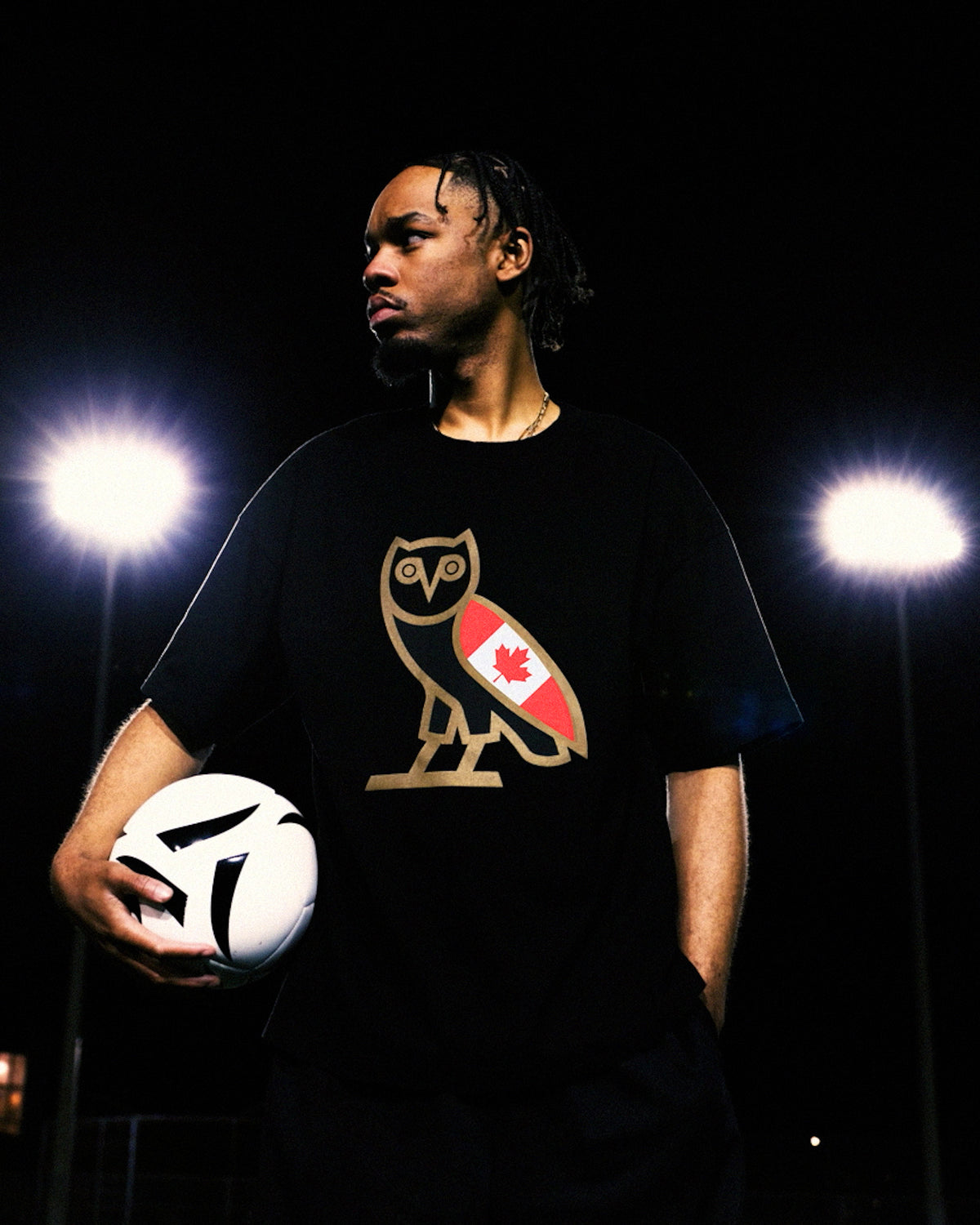 Person wearing a black October's Very Own t-shirt with a logo, holding a soccer ball on a dark background
