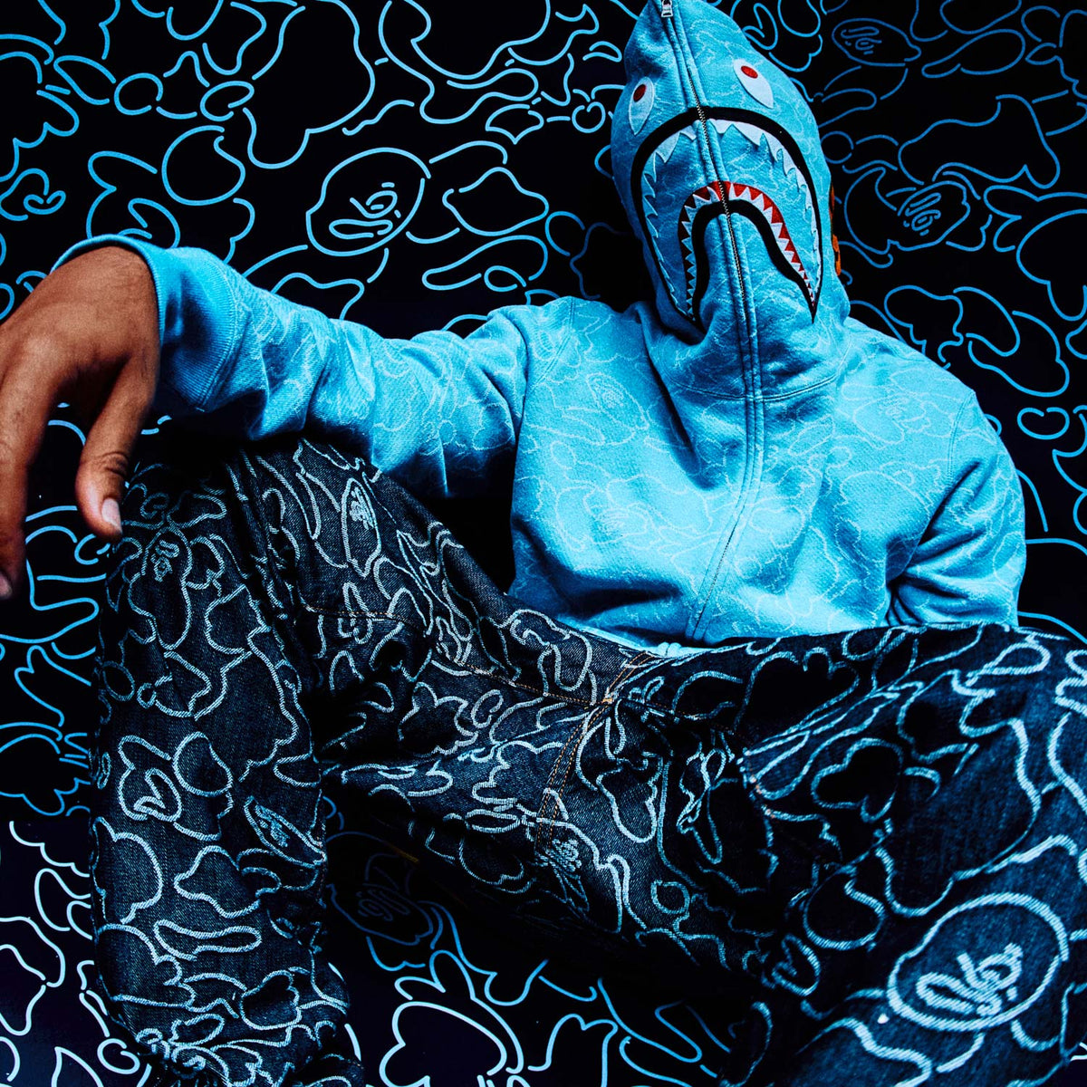 Person wearing a blue shark October's Very Own x Bape hoodie on a dark patterned background