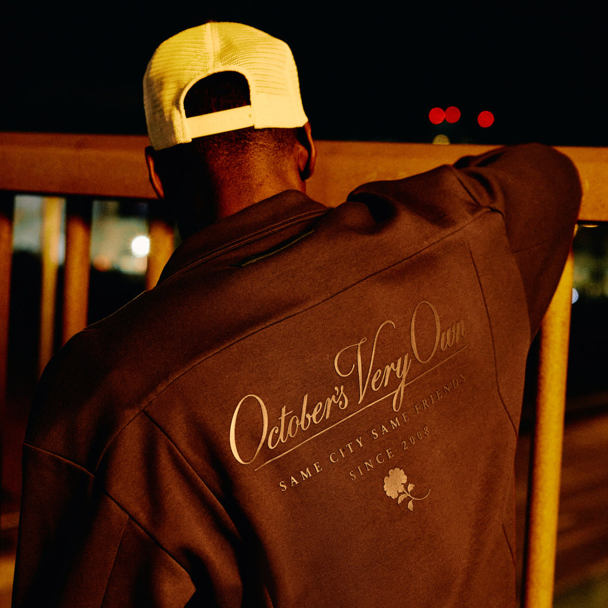 Person wearing a brown jacket with 'October's Very Own' branding, sitting on a wooden bench at night. 