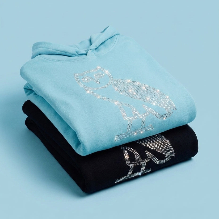Stack of light blue and black October's Very Own hoodies with rhinestone designs on a light blue background