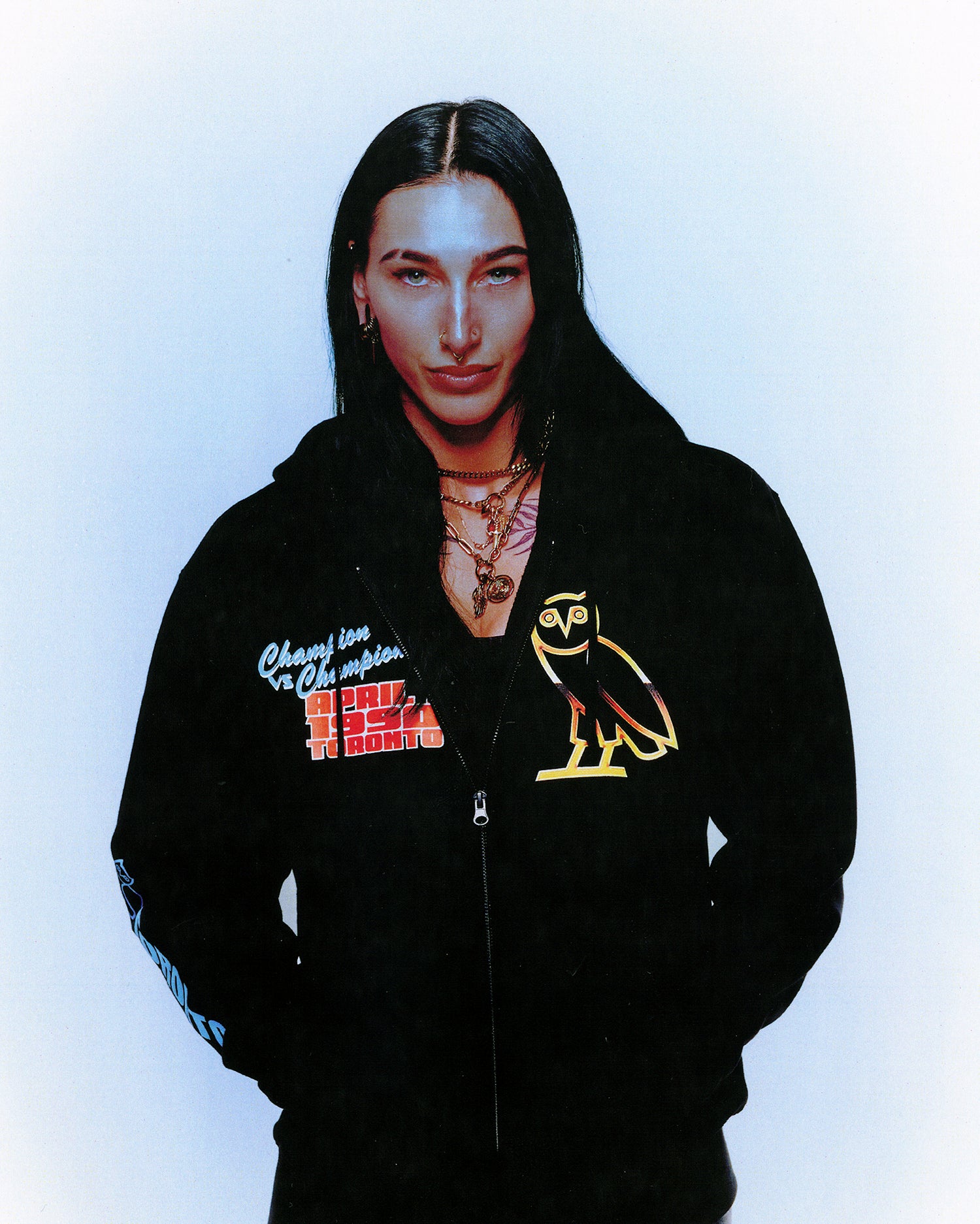 Rhea Ripley wearing a black October's Very Own x WWE hoodie with logos on a light blue background