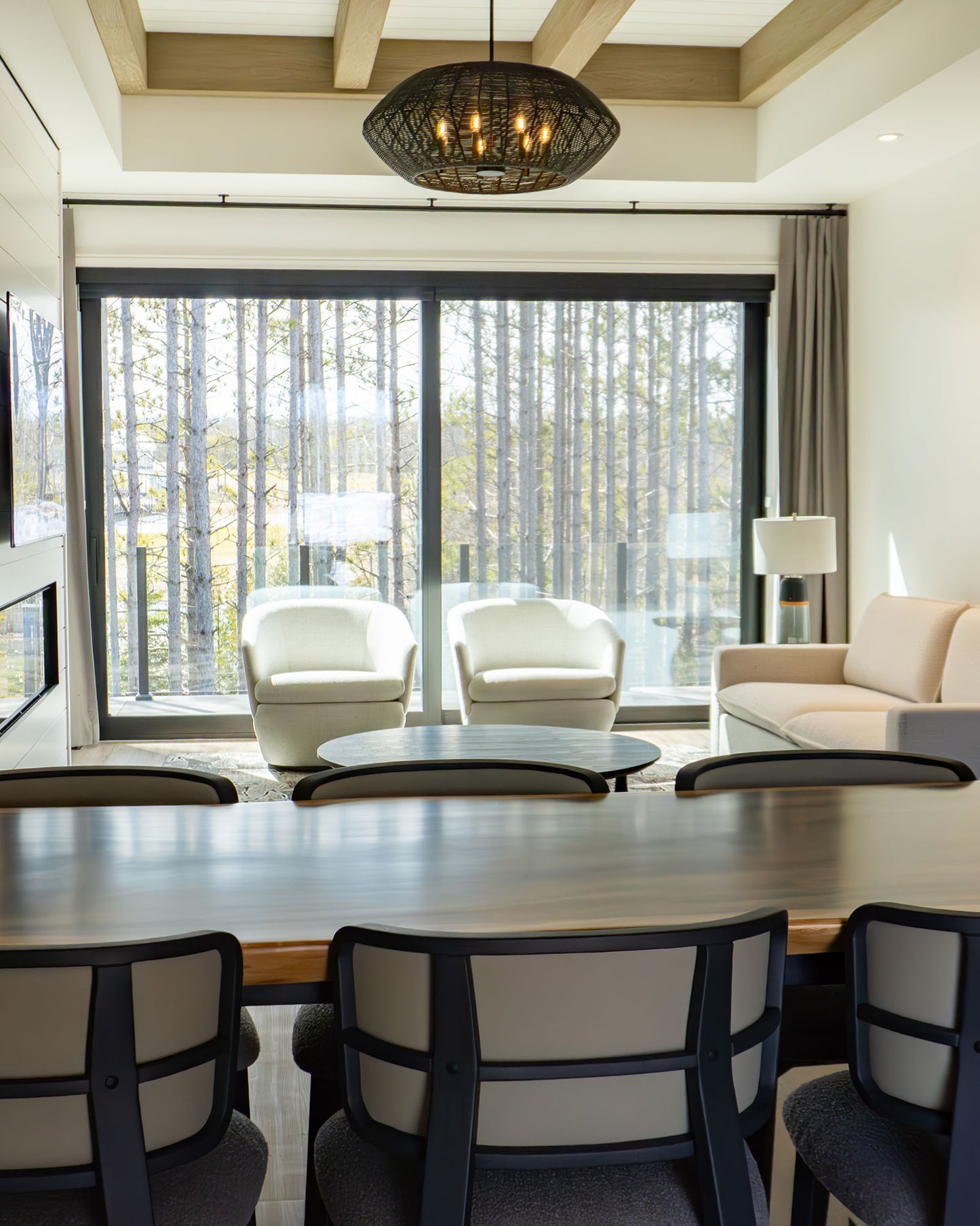 Modern lodge interior with dining table, cream armchairs, large windows overlooking forest, and pendant lighting.