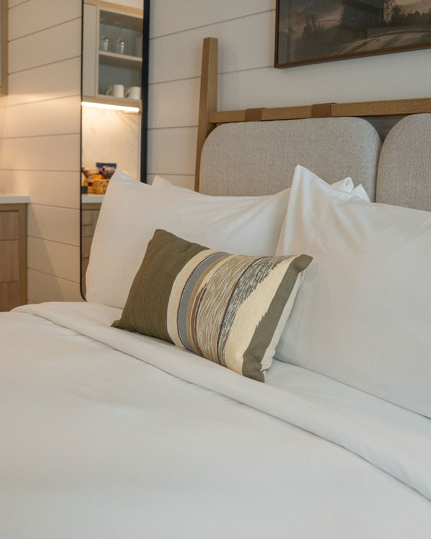 Modern hotel bedroom with wooden bed frame, white linens, striped accent pillow, and illuminated wall shelving.