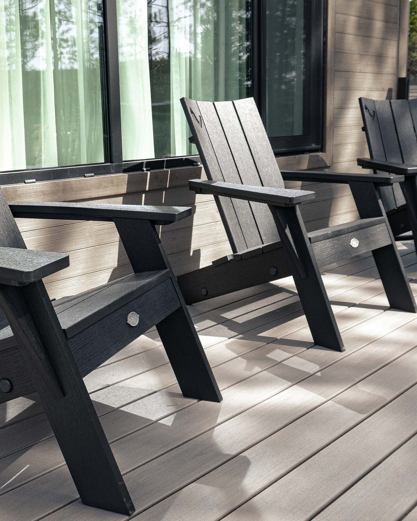 Black and gray modern adirondack chairs on a wooden deck with large windows and greenery visible behind.