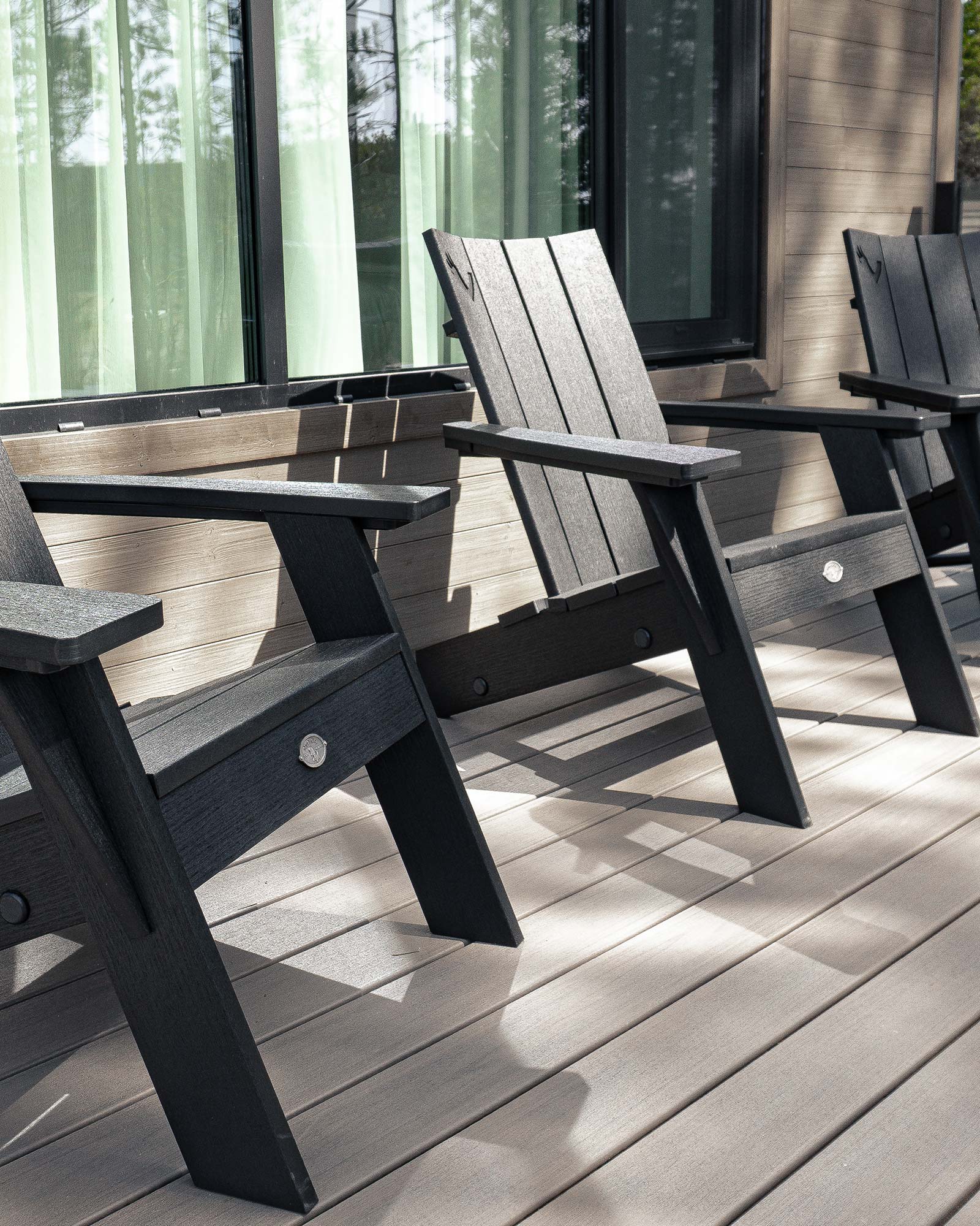 Black and gray modern adirondack chairs on a wooden deck with large windows and greenery visible behind.