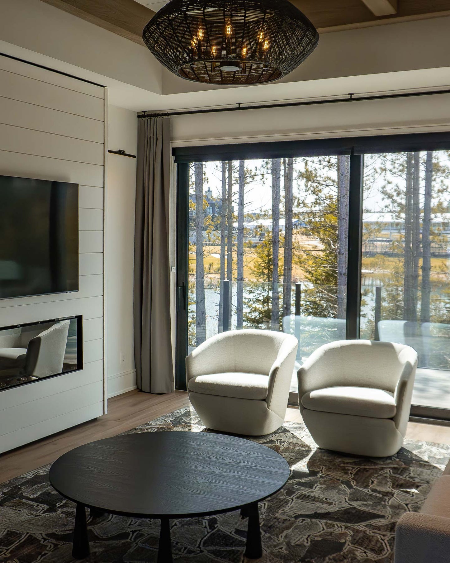 Luxury resort suite with two cream armchairs, round black table, fireplace, TV, and lake views through large windows.