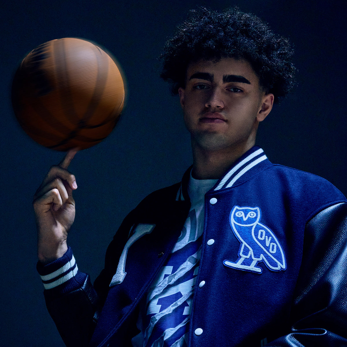 Person holding a basketball wearing a blue letterman jacket with an owl logo on a dark background wearing the October's Very Own an Kentucky Wildcats collection