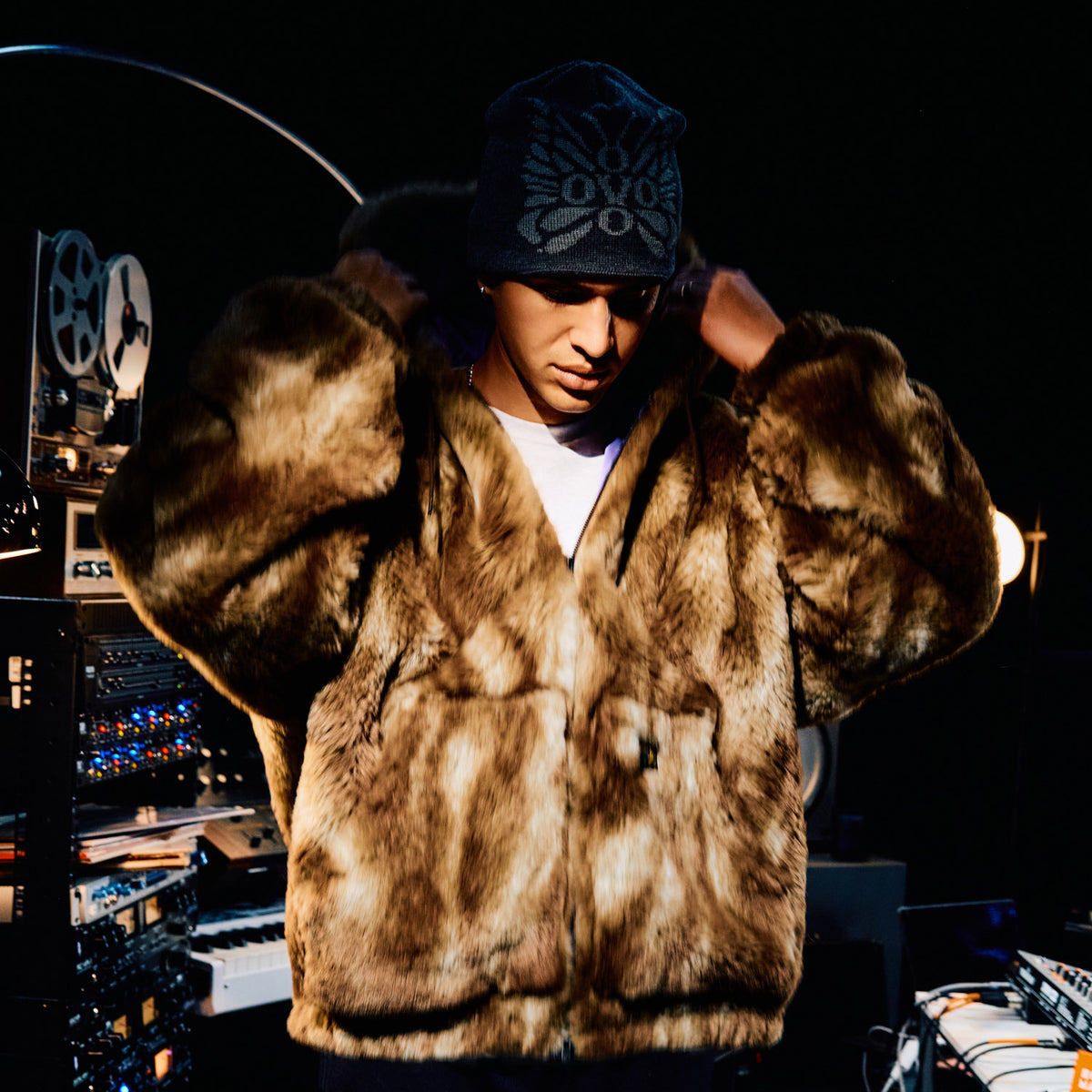 Person wearing a fur vest and cap in a dark room with equipment. October's Very Own and Needles.