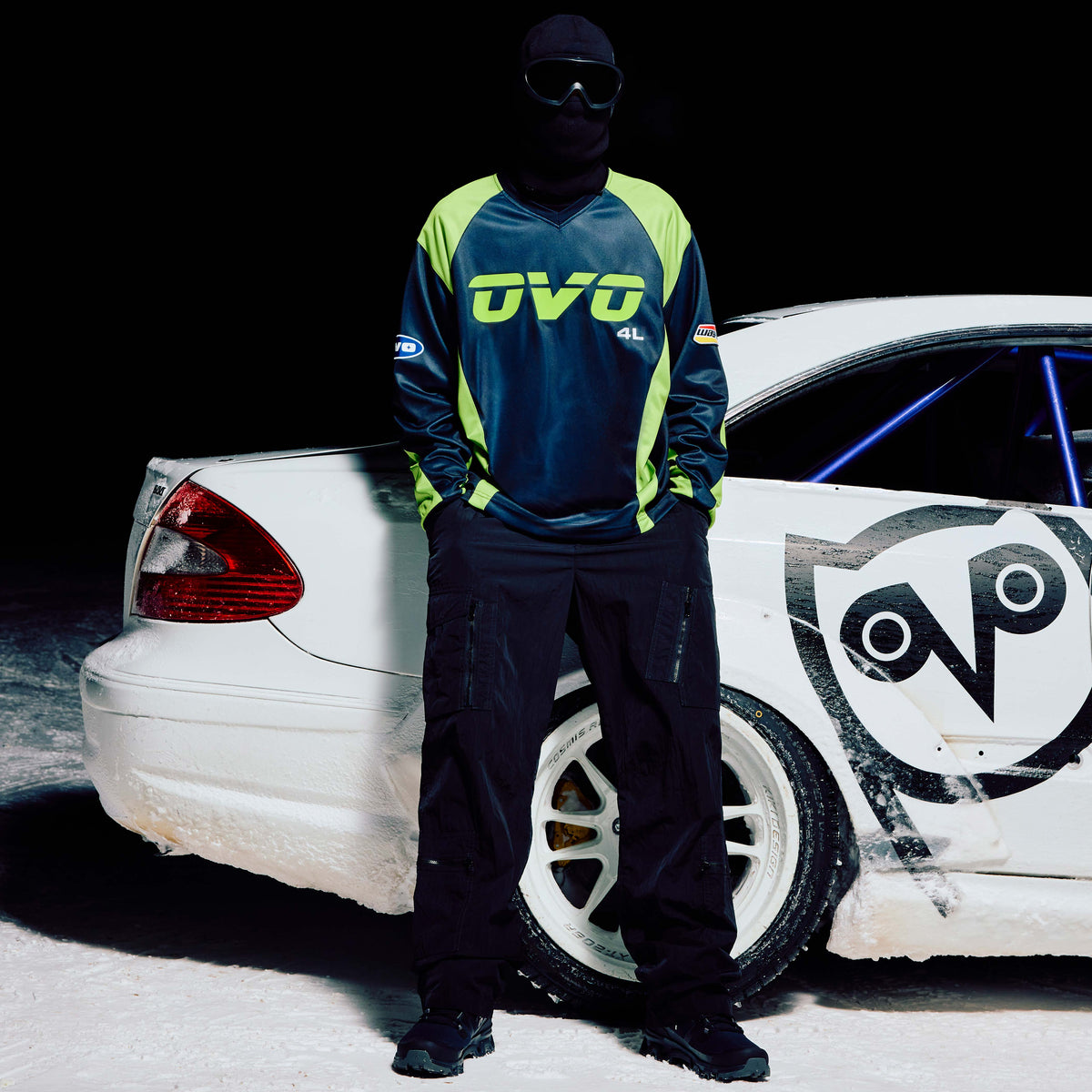 Person wearing a blue and green jacket with 'OVO' branding standing next to a white car with similar branding.