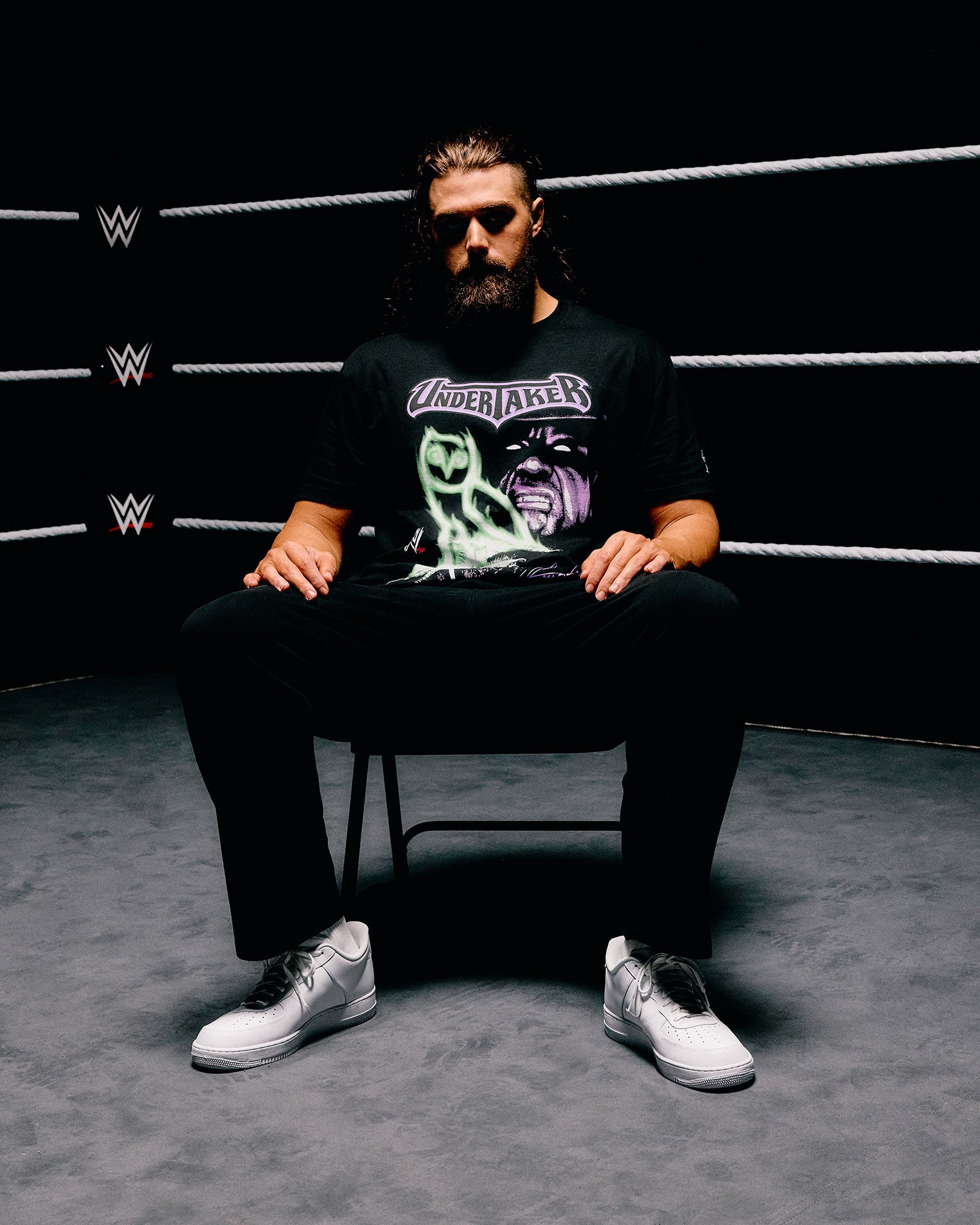 Person sitting in a chair wearing a black October's Very Own x WWE t-shirt with a graphic design, in a wrestling ring setting.
